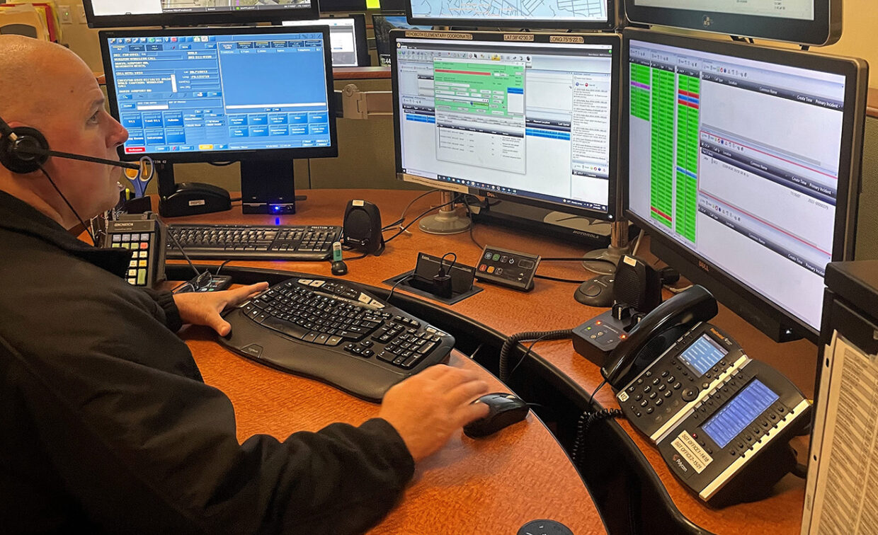 A person wearing a headset sits at a desk with multiple computer monitors displaying various information.