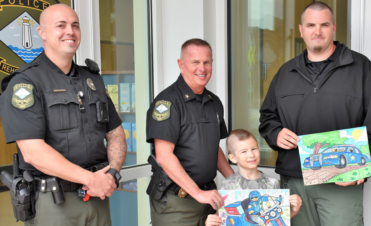 Three uniformed police officers stand with a child holding colorful drawings of police vehicles. They are in front of a building entrance with a police logo.