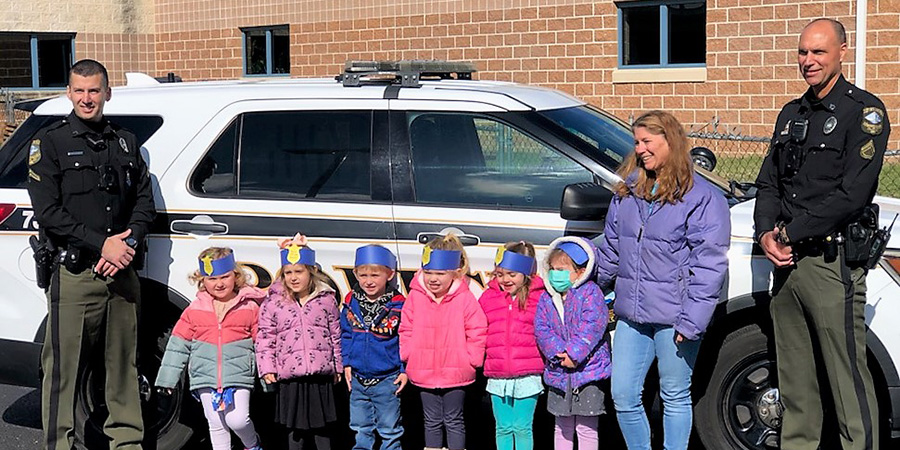 Two police officers, a woman, and six children wearing colorful hats stand in front of a police car outside a brick building.