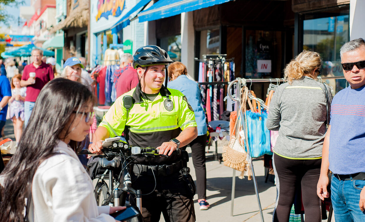 A police officer on a bike interacts with people on a busy street lined with shops and stalls.