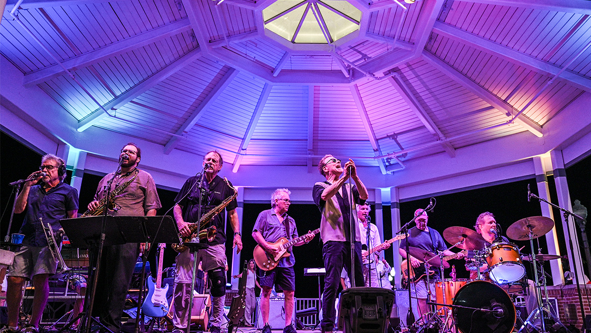 A band performs under a lit gazebo at night, with members playing saxophones, trumpets, guitars, and drums.