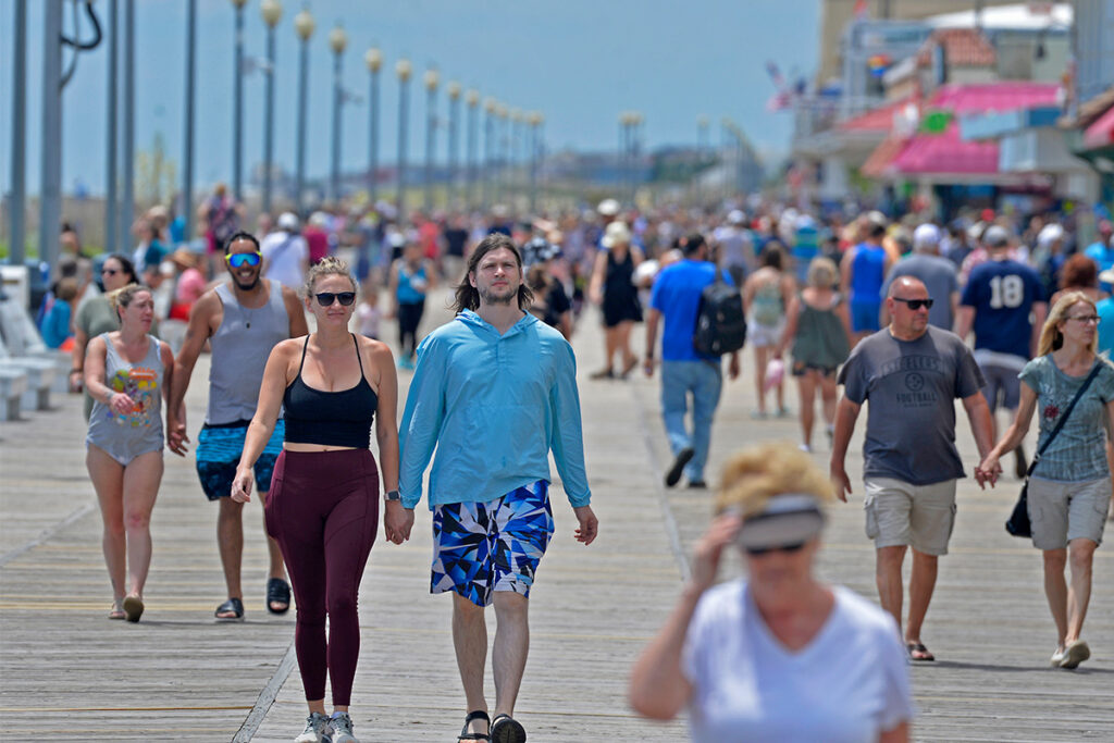 People walking along a crowded boardwalk on a sunny day, with shops and streetlights in the background.