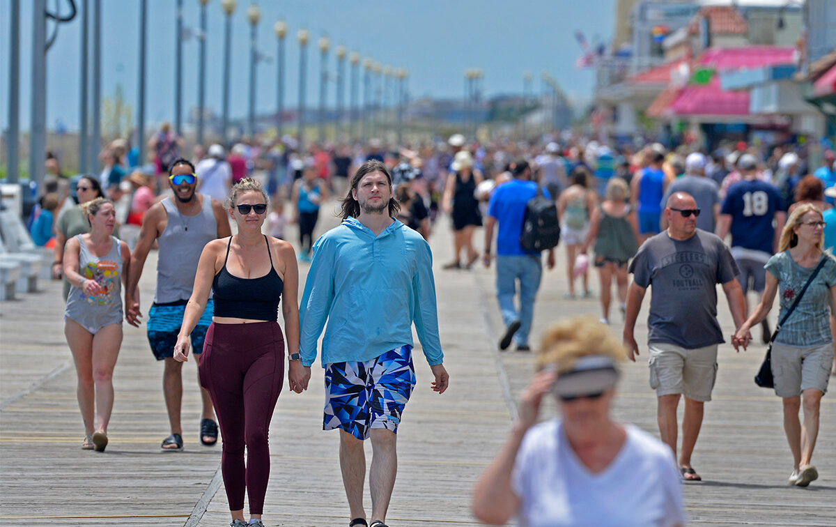 People walking along a crowded boardwalk on a sunny day, with shops and streetlights in the background.