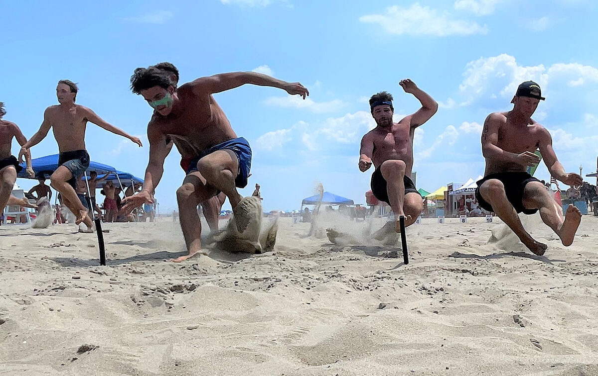 People racing on a sandy beach, jumping over obstacles with determination. Blue sky and beach tents in the background.