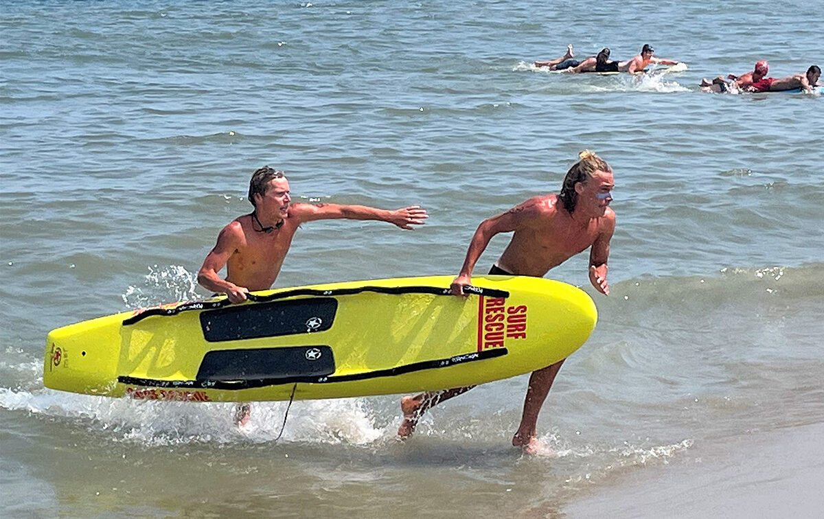 Two lifeguards carry a yellow surf rescue board out of the ocean, with other swimmers visible in the background.