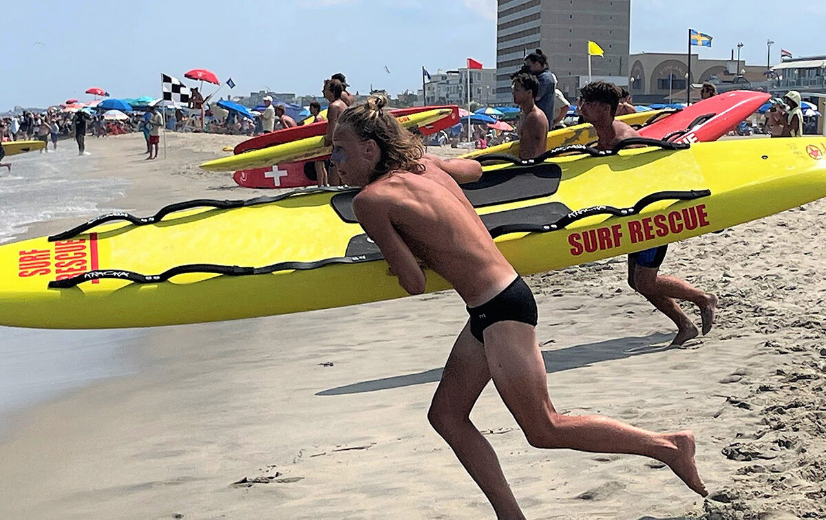 Lifeguards running with surf rescue boards across a sandy beach, with beachgoers and umbrellas in the background.