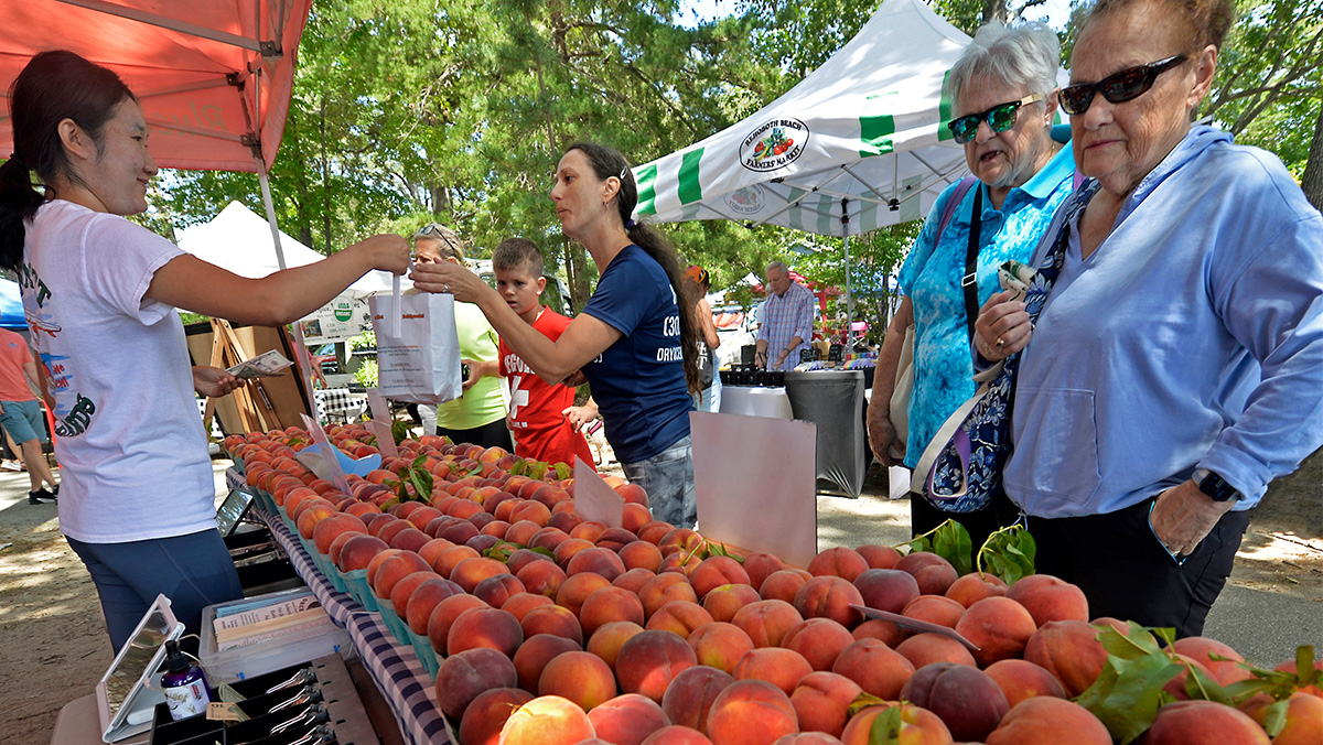 People buying peaches at an outdoor market stand under tents, with trees in the background.