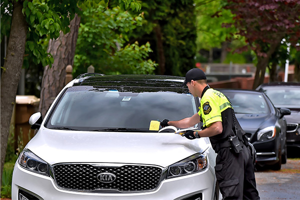 A parking enforcement officer issues a ticket to a white SUV parked on a tree-lined street.