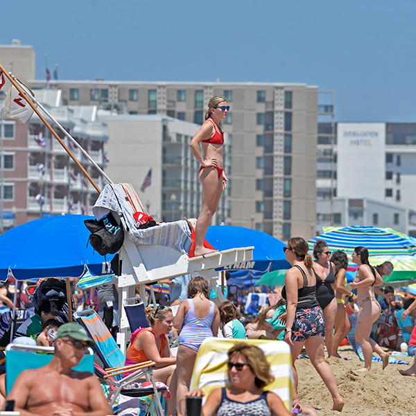 A lifeguard in a red bikini stands on a chair, overseeing a crowded beach with people under umbrellas and swimming. Background features tall buildings.