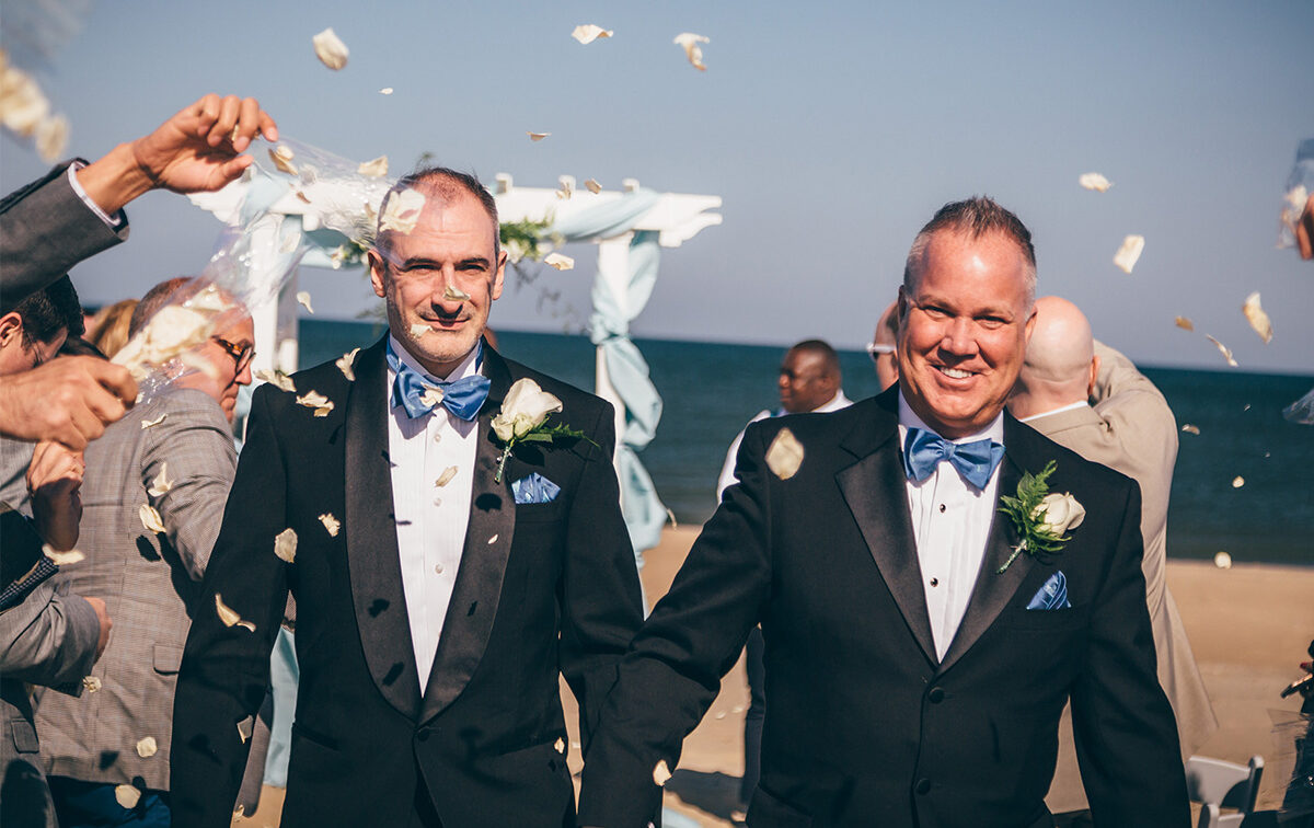 Two men in tuxedos holding hands after a wedding ceremony on a beach, with people tossing confetti around them.