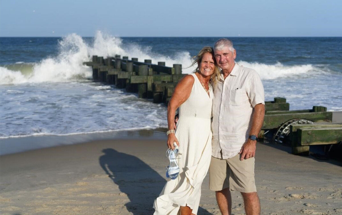 A smiling couple stands on a beach with waves crashing against a jetty in the background. The woman holds sandals in her hand.