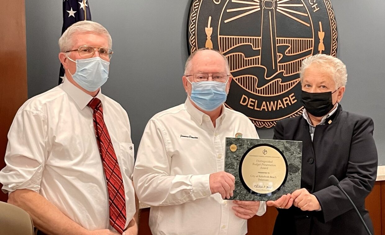 Three individuals stand in front of the Lewes, Delaware city seal; two hold an environmental sustainability award. The men wear masks; the woman is dressed in a black suit.