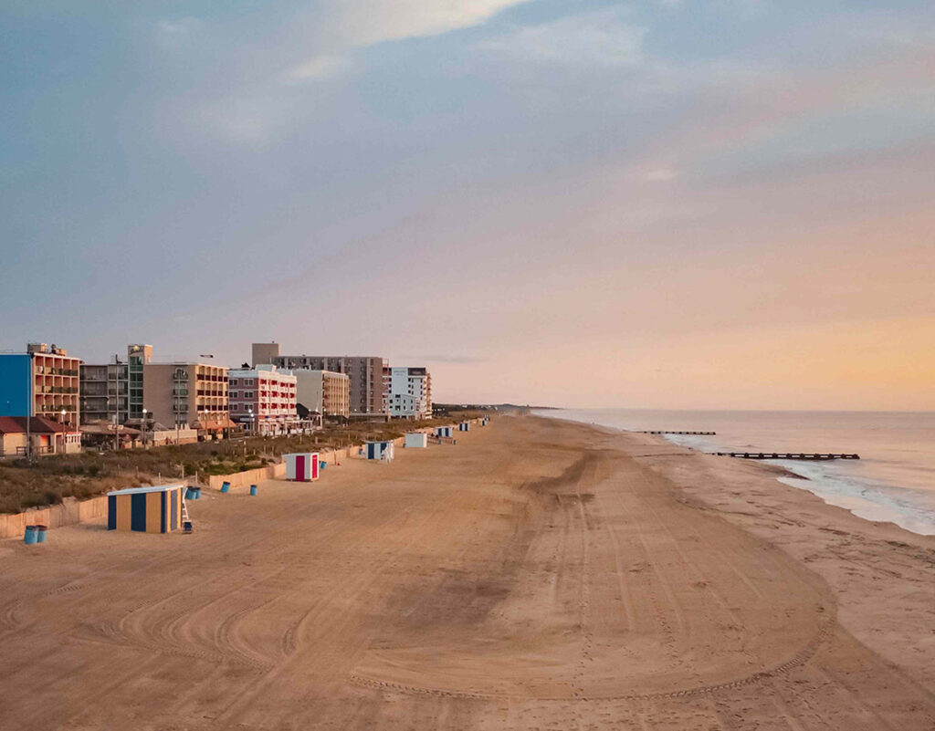 Wide sandy beach with colorful cabanas, beachfront buildings, and calm ocean under a pastel sky at sunrise.