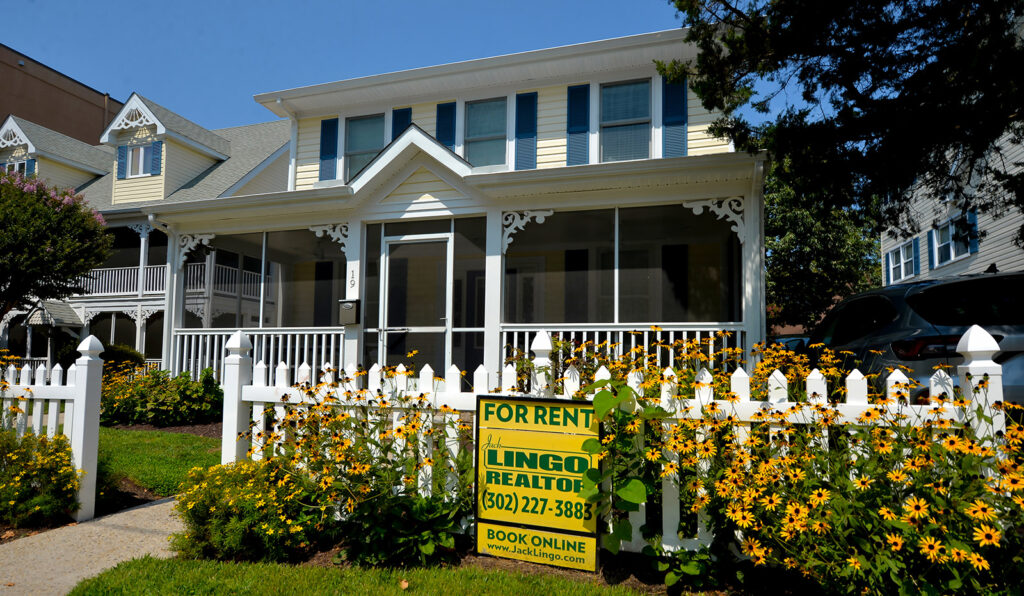 A white two-story house with a porch and picket fence is available for rent. A "For Rent" sign from Lingon Realtors is displayed in the front yard.