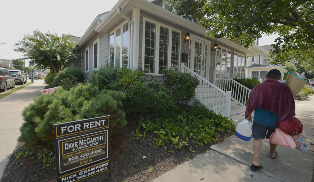 A person carrying items walks on a sidewalk past a "For Rent" sign in front of a residential house with a porch and greenery.