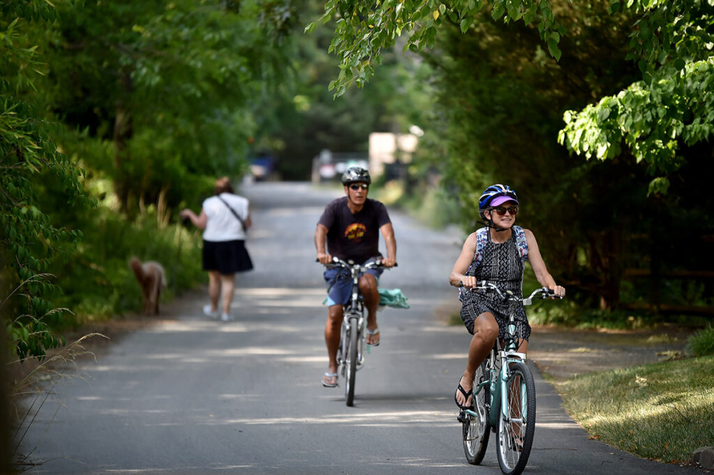 Two people ride bicycles on a shaded path. Another person walks a dog in the background.