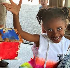 A smiling child holds a colorful painted craft with people and a child in the background.