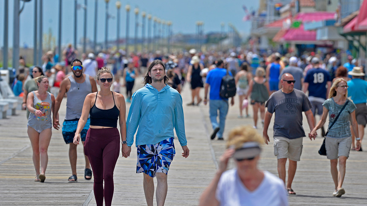 A crowded boardwalk on a sunny day, with people walking and some in beach attire.