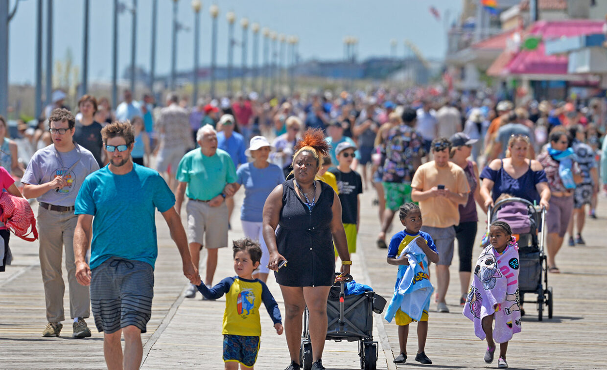 A crowded boardwalk scene with people walking, some pushing strollers, and others pulling wagons.