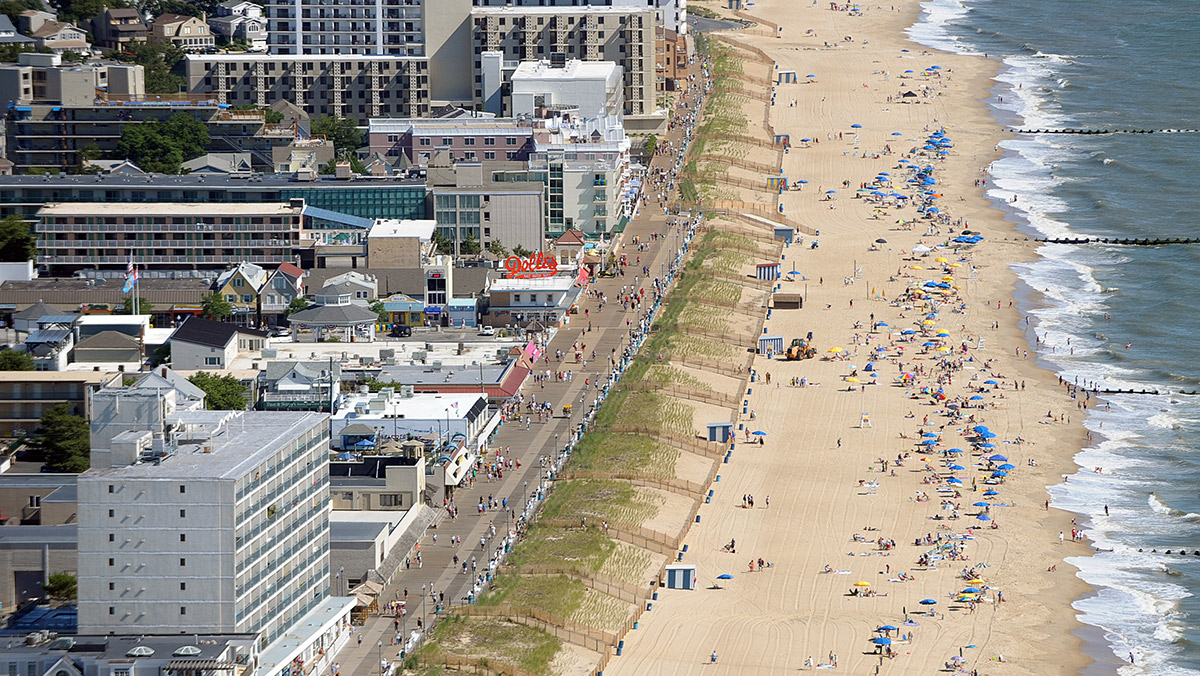 Aerial view of a bustling beach and boardwalk lined with buildings and people, with the ocean on the right and numerous beachgoers enjoying the sand and water.