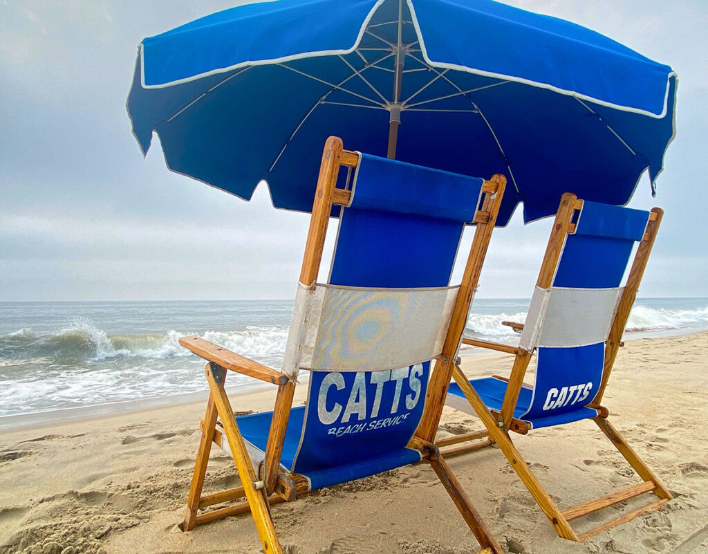 Two wooden beach chairs with blue and white fabric sit under a matching umbrella on a sandy beach, facing the ocean.