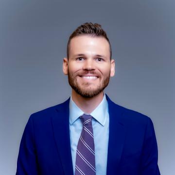 A man with short hair and a beard is wearing a blue suit, a light blue shirt, and a striped tie, standing against a plain gray background.