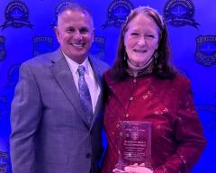 Two people are standing side by side against a blue backdrop with logos. The woman on the right is holding an award. Both are smiling.