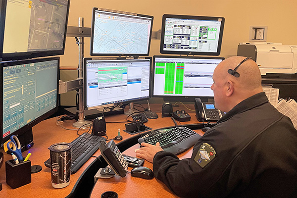 A person in a dark uniform sits at a desk with multiple computer monitors displaying maps and data.