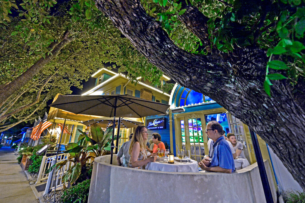 A couple dines outdoors at a restaurant under a large tree at night. The restaurant is brightly lit, and other diners are visible in the background.