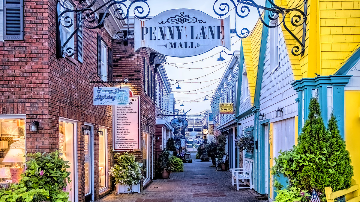 Narrow alleyway leading to Penny Lane Mall, flanked by colorful buildings and hanging lights, with plants lining the path.