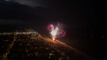 Fireworks illuminate the night sky over a beach, with a cityscape visible on the left side of the image.