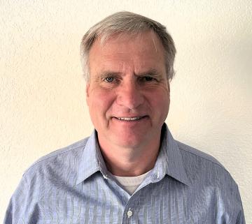 Man with short gray hair and a light blue shirt stands against a plain white background.