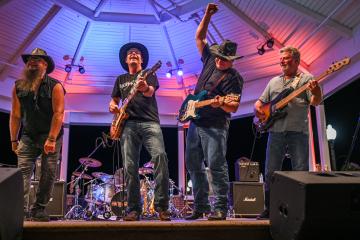 Four musicians performing on stage under a gazebo, playing guitars and wearing hats, with drums and amplifiers in the background.