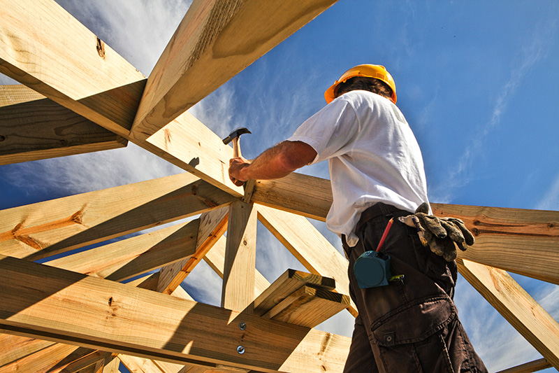 Construction worker in a hard hat hammering wood beams on a roof under a blue sky.