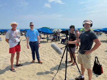 Four men stand on a sandy beach next to a microphone and tripod, with blue umbrellas and people in the background.