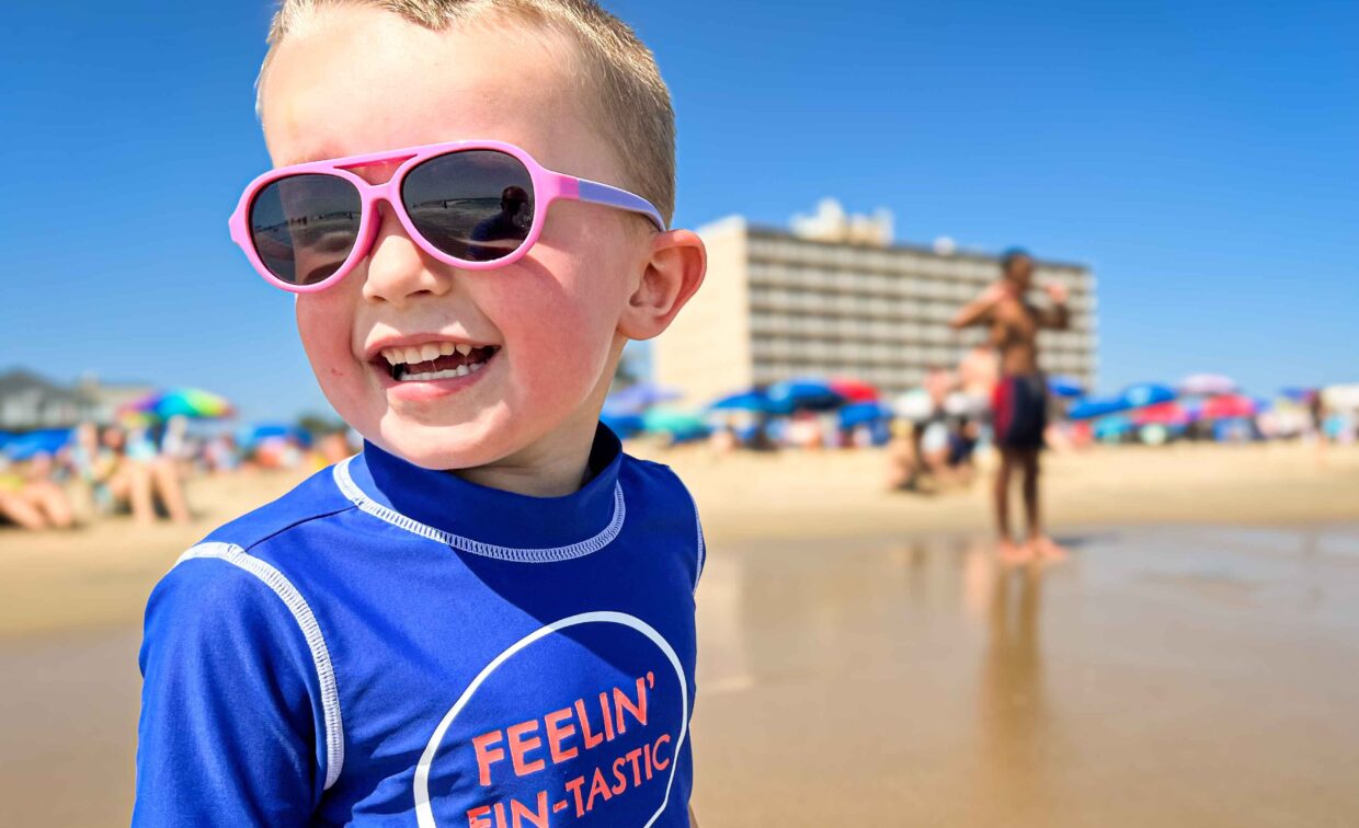 A 2024 Rehoboth Reflections-winning photo of a cute boy in pink sunglasses and a Fellin' Fintastic shirt standing on the water's edge