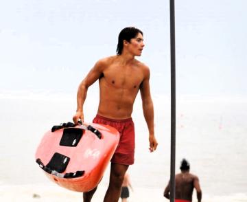 A person in red shorts carries a red surfboard on a beach with the ocean in the background.