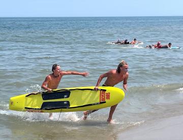 Two people carry a yellow surfboard out of the ocean while others swim in the background.