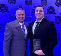 Two men smiling at an event in front of a backdrop with emblems. One man holds an award plaque. They are both wearing suits. The lighting is blue-toned.