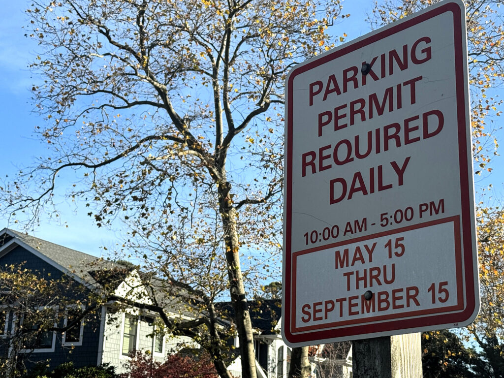 Sign indicating parking permit is required daily from May 15 to September 15, 10:00 AM to 5:00 PM. Background shows houses and trees.