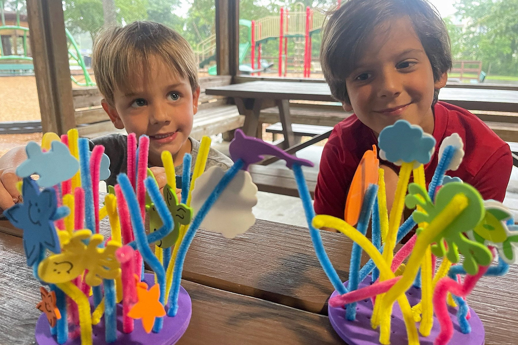 Two children smiling behind colorful pipe cleaner crafts with foam cutouts at a picnic table in an outdoor setting.