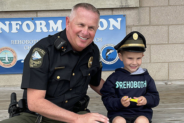 A police officer and a young child in a police hat smile for a photo. The child holds a small object. They are in front of an information booth sign.