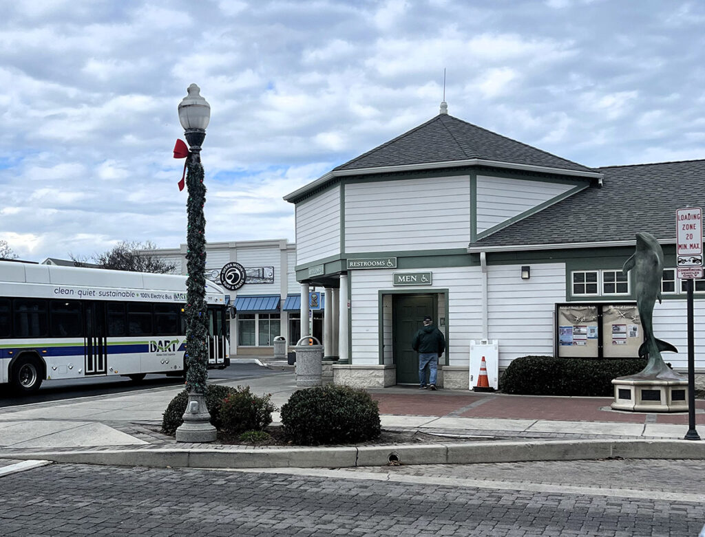 A man stands outside a freestanding restroom building near a bus stop. A bus approaches on the street. The area is decorated with a lamppost and a sculpture. Cloudy sky in the background.