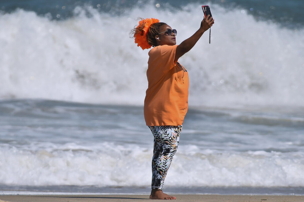 A person wearing an orange top and patterned leggings takes a selfie on a beach, with waves crashing in the background.