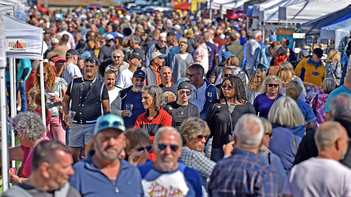 A large crowd of people walk through an outdoor market or fair, browsing various vendor stalls under white tents on a sunny day.