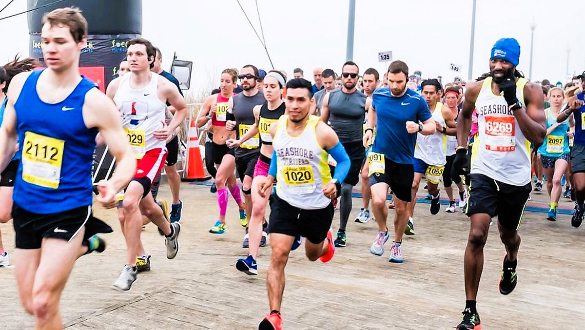 Runners in a race wearing numbered bibs, various athletic outfits, and sneakers, competing on a wooden boardwalk.