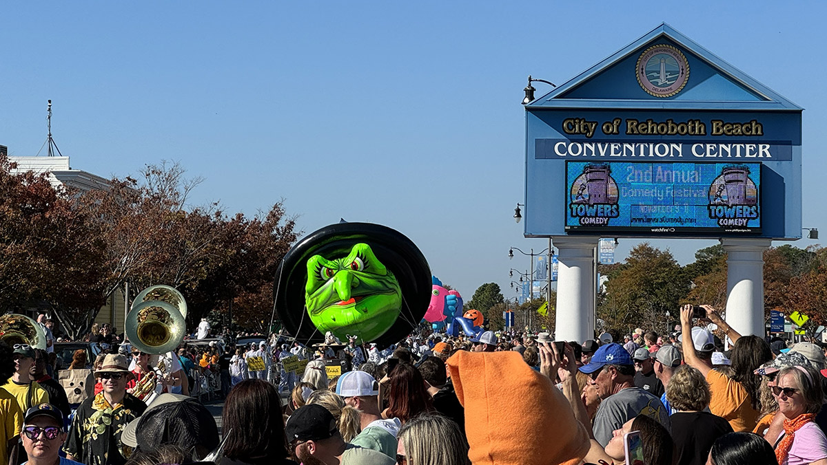 A parade with a green witch balloon passes by a crowd near the City of Rehoboth Beach Convention Center.