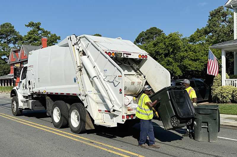 Garbage collectors empty bins into a white truck on a suburban street, with trees and a house in the background.