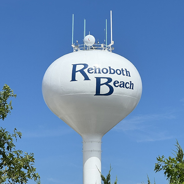 White water tower with "Rehoboth Beach" written on it, set against a clear blue sky and surrounded by green trees.